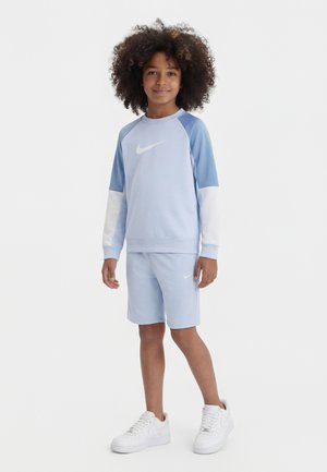 Dziecko o kręconych włosach, ubrane w jasno niebieską bluzę Nike i szorty, stojące w białych trampek na białym tle.