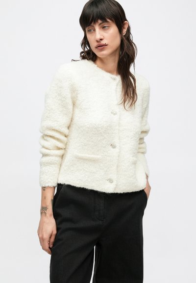 Bela jopica z mehko teksturo, okrogel izrez, sprednjimi žepi in petimi dekorativnimi gumbi. Oblečena čez črne hlače.