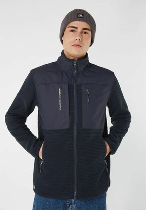 Giacca in felpa della marina con colletto alto, pannelli laterali testurizzati, due tasche sul petto con zip e tasche laterali con zip coordinate. Vestibilità casual.