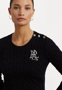 Zwarte kabelgebreide trui met een ronde hals, zilveren schouderknopen en een geborduurd logo op de borst. Gladde textuur.