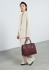 Burgundy leather handbag with top handles, structured shape, subtle grain texture, and gold hardware accents, held by a person in a light suit.