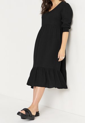 Kobieta ubrana w czarną sukienkę midi z bufkami na rękawach i falbaną na dole, w połączeniu z czarnymi sandałami wsuwanymi, stojąca na białym tle.