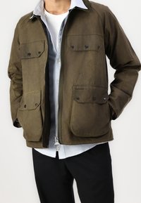 Mann trägt eine olivgrüne Barbour-Jacke mit vier Klapptaschen, ein hellblaues Hemd, ein weißes T-Shirt und schwarze Hosen, die Hände in den Taschen.