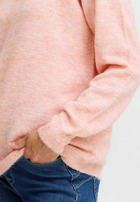 Maglione a maglia rosa chiaro con una texture morbida, polsini e orlo a coste. Indossato sopra jeans in denim blu, con una mano nella tasca.