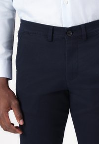 Marinblå bomullsbukser med en slät yta, framknäppning med knapp och en smal passform. Har ett subtilt diamantmönster.