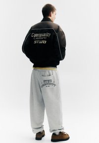 Jachetă bomber neagră cu bordură albă și text auriu pe spate, asortată cu pantaloni de trening gri cu logo imprimat, purtată cu pantofi maro.