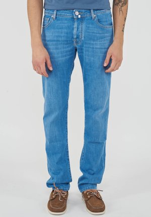 Uomo che indossa jeans azzurri chiari a gamba dritta e scarpe da barca marroni, in piedi davanti a uno sfondo semplice.