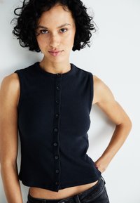 Débardeur côtelé bleu marine sans manches avec un col rond et une fermeture à boutons sur le devant avec des boutons noirs. Texture lisse et design court.