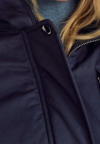 Veste matelassée bleu marine avec une fermeture éclair, des accents de boutons et une texture douce. Dispose d'un col montant et d'un sous-pull gris contrastant.