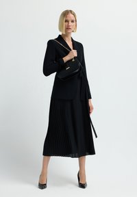 Frau in schwarzem Blazer und plissiertem Midirock, die eine schwarze Handtasche mit goldener Kettenriemen hält, in schwarzen High Heels stehend vor weißem Hintergrund.