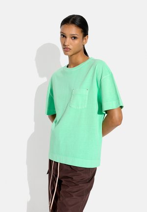 Kobieta o ciemnych włosach, ubrana w miętową koszulkę z krótkim rękawem i kieszenią z przodu, brązowe spodnie, stojąca na tle jasnego, jednolitego tła.