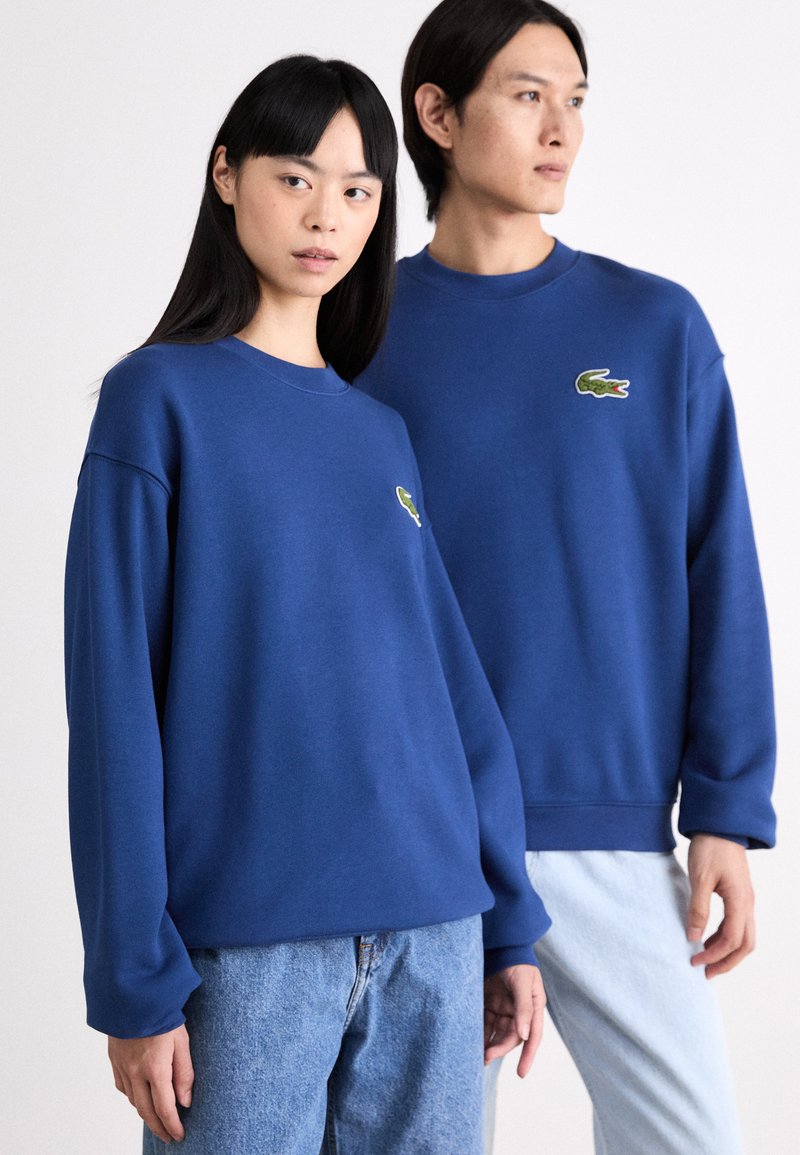 Sudaderas azules de corte relajado, con cuellos redondos y el logo de Lacoste bordado en verde en el lado izquierdo del pecho. Pantalones de mezclilla.