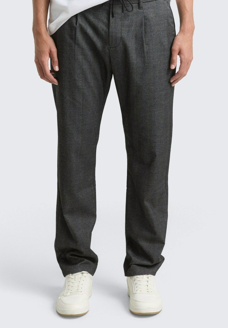 Mann trägt dunkelgraue, locker sitzende Hose und weiße Turnschuhe, steht mit entspannten Händen seitlich an einem einfarbigen Hintergrund.