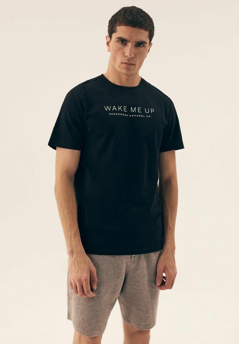 Czarna bawełniana koszulka z krótkim rękawem, z białym napisem "OBUDŹ MNIE" oraz "HENDERSON APPAREL CO." na środku. Połączona z szarymi szortami.