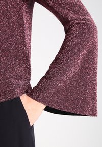 Top a maniche lunghe con maniche a campana, realizzato in un tessuto bordeaux scintillante. Presenta un design aderente e una texture liscia, abbinato a materiale nero.