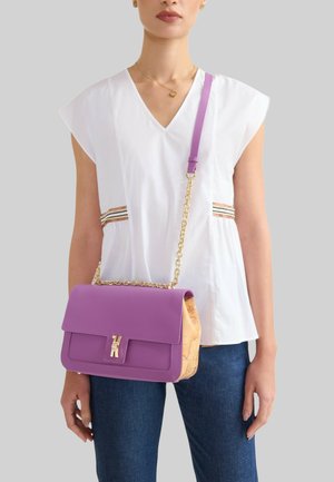 Donna che indossa una canotta bianca e jeans blu, con una borsa a tracolla viola e beige con cinghia a catena dorata.