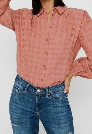 Vrouw met een gestructureerd, roze overhemd met lange mouwen, ingestopt in een blauwe spijkerbroek, met de ene hand op de heup en de andere omhoog geheven.