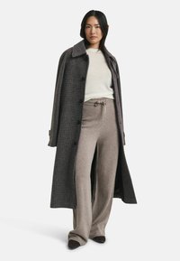 Manteau long à carreaux en gris foncé avec un grand col et des boutons, superposé sur un pull léger et un pantalon large beige. Texture lisse sur l'ensemble.