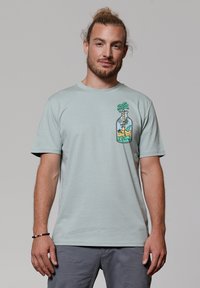 T-shirt en coton vert clair avec un col rond, présentant un graphique coloré d'une bouteille avec un palmier et une plage de sable sur le côté gauche.