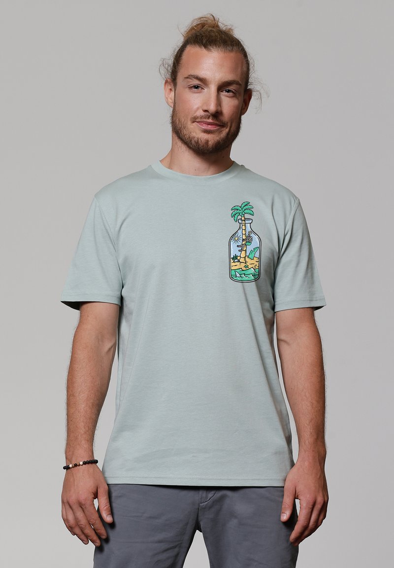T-shirt en coton vert clair avec un col rond, présentant un graphique coloré d'une bouteille avec un palmier et une plage de sable sur le côté gauche.