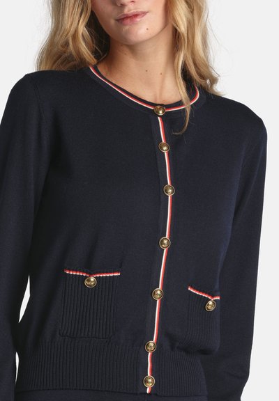 Marineblaue Strickjacke mit goldenen Knöpfen, gerippten Taschen und kontrastierendem rot-weiß gestreiftem Besatz entlang des Halsausschnitts und der Knopfleiste.