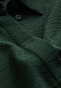 Zielona tkanina o fakturze z miękkim układem, wyposażona w dwa czarne guziki i kołnierz, ukazująca subtelne różnice w splocie i głębi.