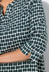 Groene en witte blouse met een patroon, V-hals en driekwart mouwen. De stof heeft afgeronde vierkantige vormen en een zachte textuur.
