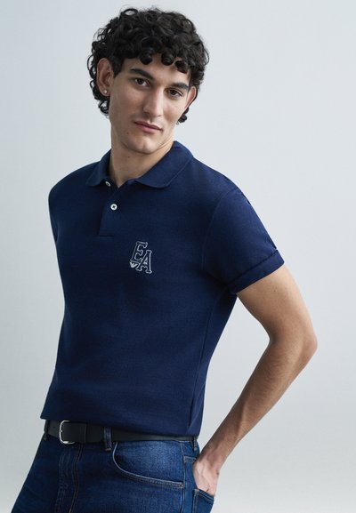 Polo bleu marine en tissu texturé, avec un col et un petit logo EA sur le côté gauche de la poitrine. Associé à un jean en denim bleu et une ceinture noire.
