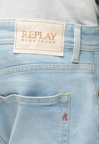 Ανοιχτό μπλε τζιν παντελόνι με λευκή δερμάτινη ετικέτα με την επιγραφή "REPLAY BLUE JEANS" ραμμένη πάνω από την πίσω τσέπη.