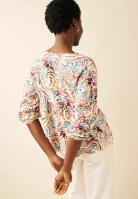 Person med kort krøllet hår og guld hoops øreringe iført en multicolor paisley bluse og hvide bukser, hænderne lagt sammen bag ryggen.