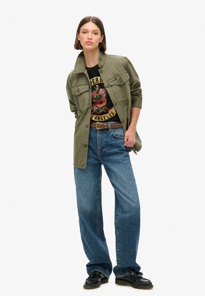 Oliwkowa kurtka robocza na czarnej koszulce z grafiką, do tego jasnoniebieskie szerokie jeansy i czarne buty, stojący na białym tle.