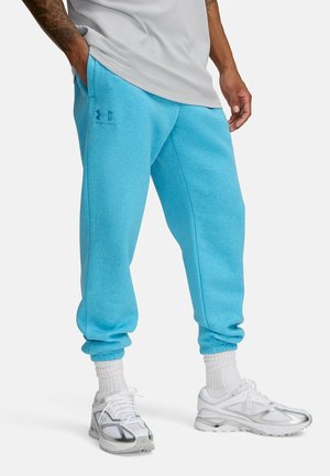 Man draagt lichtblauwe Under Armour joggingbroek, witte sokken en witte hardloopschoenen, staand met één hand in de zak.