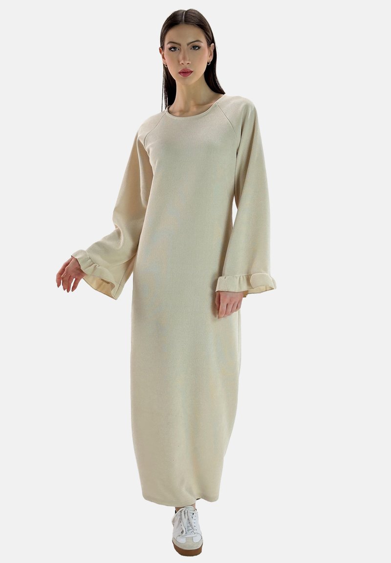 Vestido beige de manga larga con textura acanalada y puños con volantes, que presenta un escote redondo y un corte recto, combinado con zapatillas blancas.