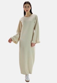 Vestido largo color crema, acanalado, con mangas largas y puños con volantes, combinado con zapatillas blancas, con una silueta relajada y recta.