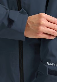 Marineblaue wasserdichte Jacke mit glatter Textur, ausgestattet mit einem Reißverschluss und Klettverschluss-Riemen an den Bündchen. "TEXPOR"-Logo am Bündchen.