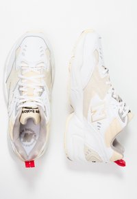 Chaussures de sport blanches et beiges avec tige en maille et synthétique, semelle en caoutchouc, logo "N" bien visible et languette rouge au talon.