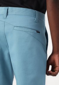 Light blue pants made of cotton, featuring a slim fit, subtle stitching details, and a back pocket with a small black logo tag.