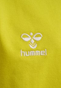 Żółta bawełniana tkanina z białym haftowanym logo w stylizowanym wzorze nad słowem "hummel" napisanym małymi literami.