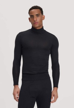 Mann trägt einen eng anliegenden schwarzen Rollkragenpullover und passende schwarze Hose, steht vor einem schlichten hellen Hintergrund.