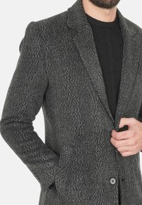 Cappotto grigio a spina di pesce con colletto a punta, tessuto testurizzato, due tasche frontali e chiusura a un solo bottone, indossato sopra un maglione nero.