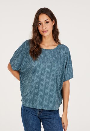 Haut teal à manches courtes avec un motif en chevrons texturé, décolleté rond et coupe ample et décontractée. Assorti à un jean bleu.