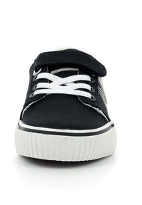 Zapatilla de lona negra con suela de goma blanca, con cordones blancos y una tira de velcro. Forma redondeada con una puntera texturizada.