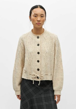 Femme portant un cardigan beige en maille torsadée boutonné, à manches longues, avec un cordon noué à la taille, associé à une jupe écossaise foncée.