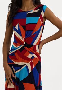 Mujer con un vestido sin mangas con un estampado geométrico abstracto en rojo, azul, negro, naranja y beige, que presenta un detalle de nudo retorcido en la cintura.