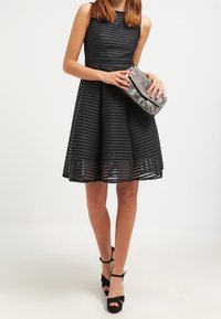 Robe noire sans manches avec des rayures horizontales texturées, jupe évasée et ourlet transparent. Associée à une pochette métallique et des chaussures à talons ouverts noires.