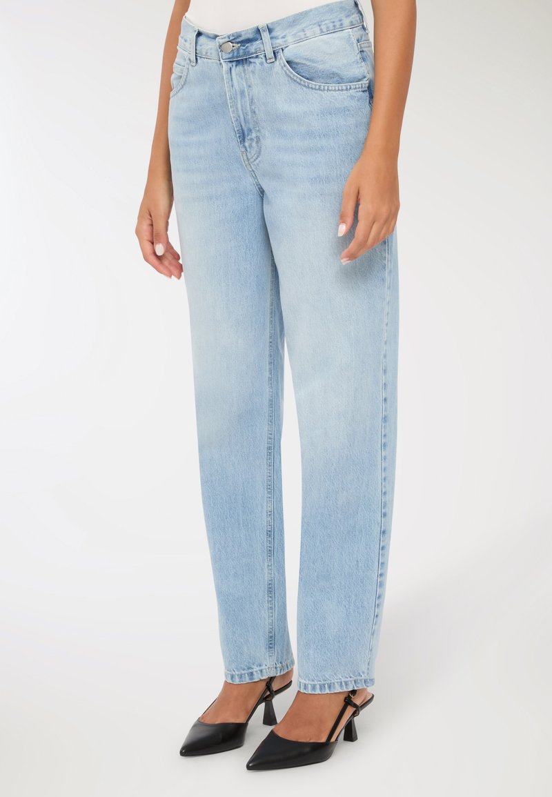 Donna che indossa jeans dritti azzurri e tacchi a punta neri, in piedi contro uno sfondo bianco.