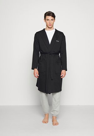 Robe de bain en coton noir avec un col châle, une taille ceinturée et deux poches avant, assortie à un pantalon de survêtement gris et pieds nus.