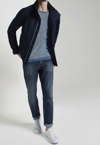 Giacca blu scuro con chiusura a zip e bottoni automatici; indossata sopra un maglione blu testurizzato. Jeans blu sbiaditi e sneakers bianche completano l'outfit.