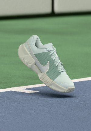 Lysegrøn og hvid Nike-sneaker med tyk sål svæver over en blå og grøn tennisbane nær grænsen.