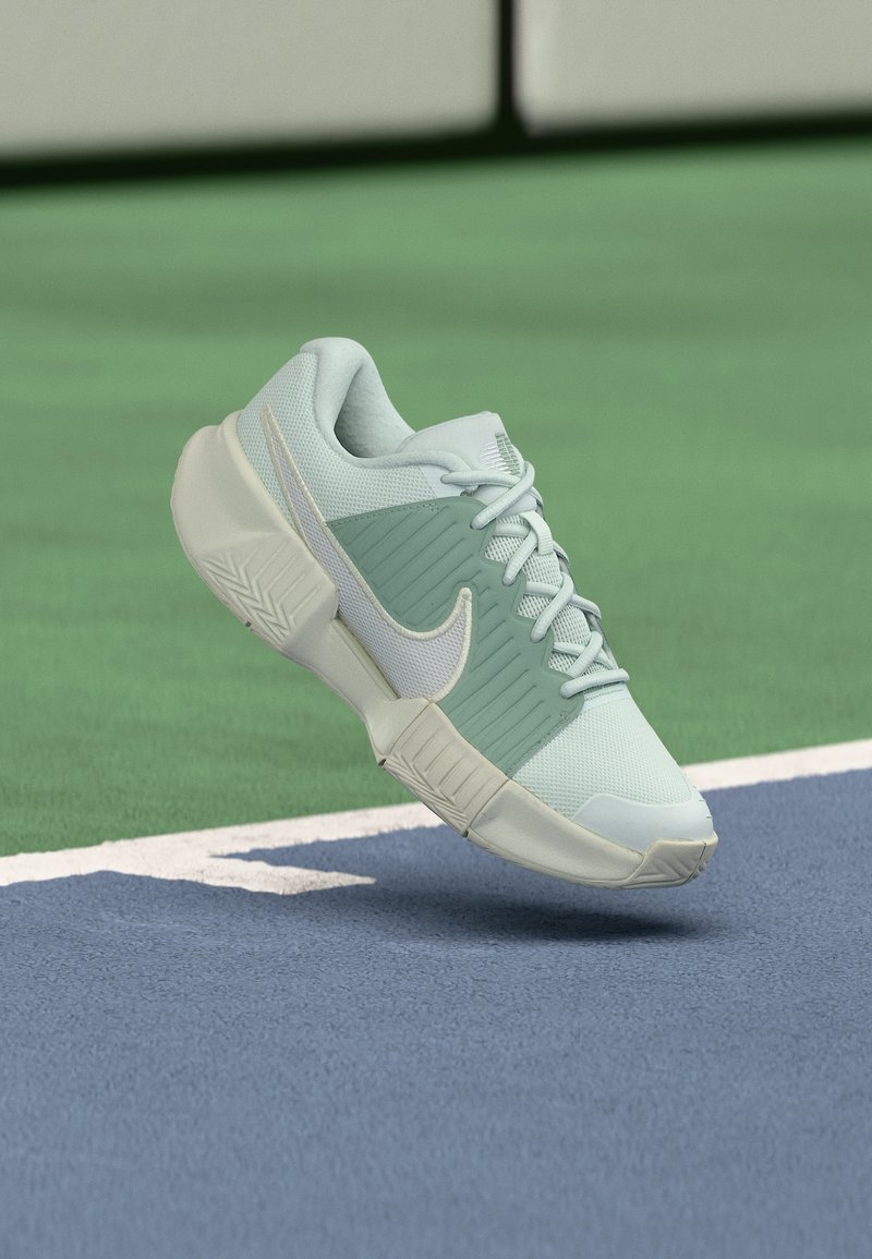 Ανοιχτοπράσινο και άσπρο αθλητικό παπούτσι Nike με παχιά σόλα αιωρείται πάνω από την μπλε και πράσινη επιφάνεια γηπέδου τένις κοντά στη γραμμή ορίων.